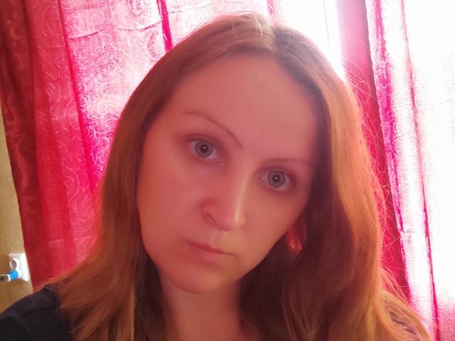 MillaOne has pale skin, red hair, and a serious expression with her brown eyes looking straight at the camera. She's wearing a black top, and the red curtains are in background.