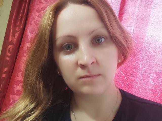 MillaOne has pale skin, blue eyes, and light brown hair. She's wearing a black shirt, looking serious, with a slight smile. Her small tits are barely visible.