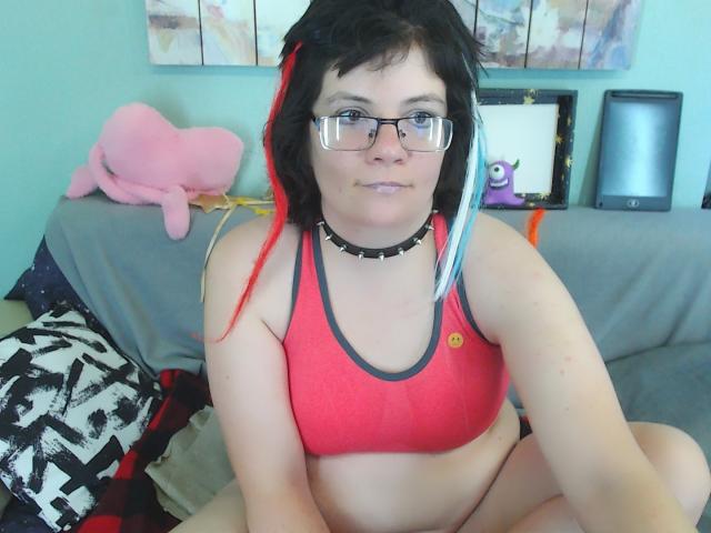 EverlyRays has short black hair with red and blue highlights, wearing glasses, a red sports bra, and a black choker, sitting with legs crossed, showing her tits.