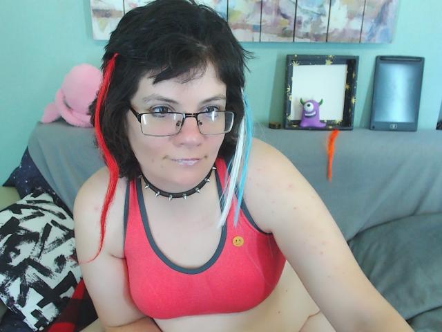 EverlyRays with short black and red-blue-dyed hair, glasses, and a red sports bra, showing her tits, wearing a spiked choker, sitting on a gray couch.