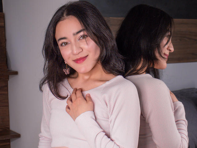 VioletteDoreens with dark hair, pink ribbed sweater, and pink earrings, smiling softly, hand on chest, reflected in mirror, showing her back and long hair.