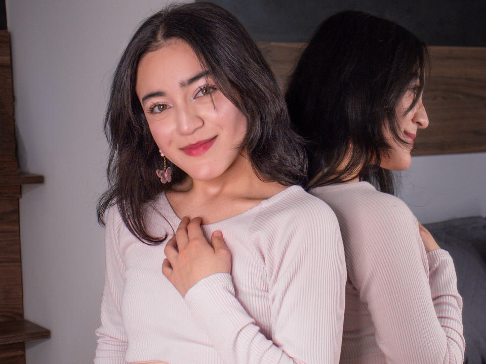 VioletteDoreens with dark hair, pink ribbed sweater, and pink earrings, smiling softly, hand on chest, reflected in mirror, showing her back and long hair.
