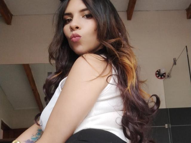 VioletaStone with long, wavy dark brown and orange hair, pouty lips, white tank top, black skirt, tattoo on left arm, looking over shoulder, wooden ceiling beams.