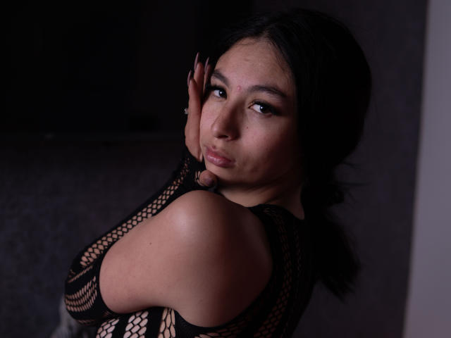 CamilleRios with dark hair, looking back, wearing a black fishnet top, exposing her bare shoulder and small, perky tits, hands on her cheek, soft smile, dark background.