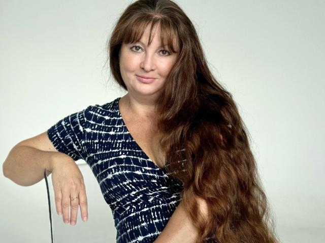 OVanessaHotly has long, wavy brown hair, wearing a blue and white patterned dress, sitting with one arm resting on a chair, smiling softly. Her tits are small.