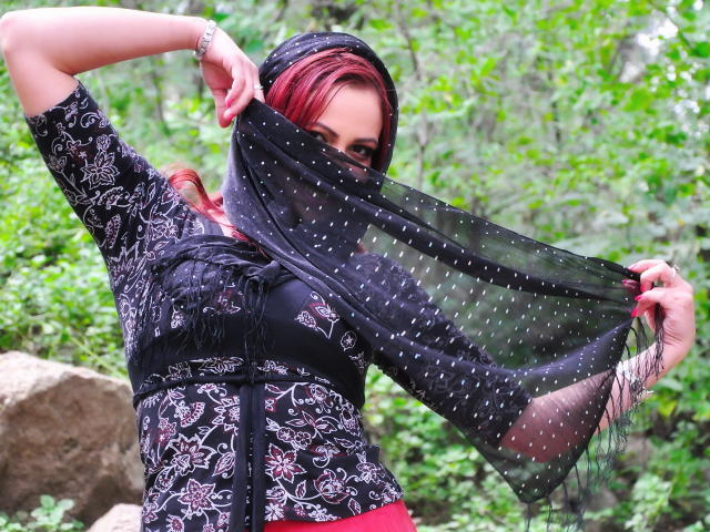 LadyHellena with red hair, black patterned top, and black polka-dot sheer scarf covering her face, holding scarf up, in a forest setting, showing her tits.