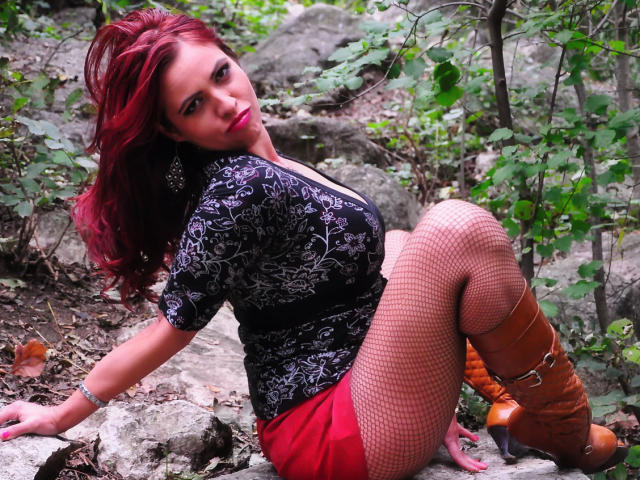 LadyHellena with red hair, black floral top, red skirt, fishnet stockings, and brown boots, sitting on rocks, looking back with a slightly open mouth, showing her ass.