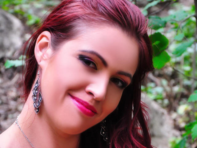 LadyHellena has red hair, pink lipstick, black eyeliner, and silver drop earrings. She's smiling with a slight tilt, showcasing her cute face and shiny skin.