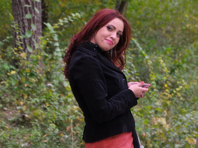 LadyHellena with red hair, black jacket, and orange skirt, holding phone, standing in forest, looking over shoulder, smiling, showing small tits, cute, sexy.