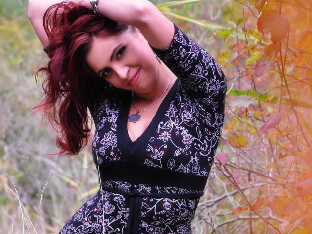 LadyHellena with red hair, wearing a black floral dress, arms raised, smiling, showing her tits, and a tattoo under her neck, standing in a grassy, leafy area.