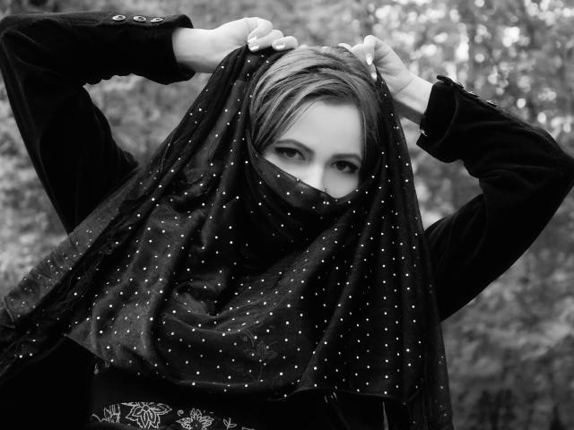 Il volto di LadyHellena è parzialmente coperto da una sciarpa nera a pois, e le sue mani la tengono sollevata, rivelando i suoi occhi. Indossa una giacca scura a maniche lunghe. Fotografia in bianco e nero..