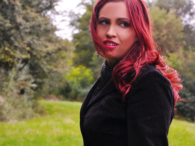 LadyHellena with red hair, black dress, and pink lipstick, smiling at the camera, showing her tits and ass in the foreground, standing in a grassy outdoor setting.