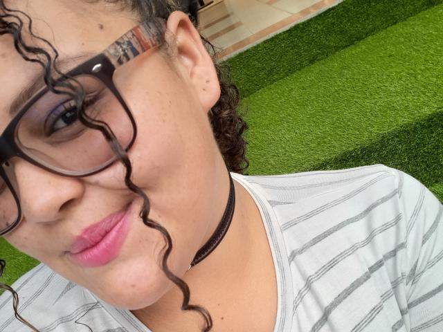 MelannieCarter has curly black hair, glasses, pink lipstick, and a black choker, wearing a striped shirt, with a green grassy background. Her tits are not visible.