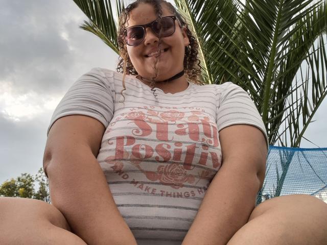 Melannie Carter mit lockigem Haar, die eine Sonnenbrille, ein weiß gestreiftes Hemd und eine Kette trägt, sitzt mit gespreizten Beinen, wobei ihre dicken Oberschenkel sichtbar sind, und lächelt. Im Hintergrund sind Palmenblätter zu sehen..