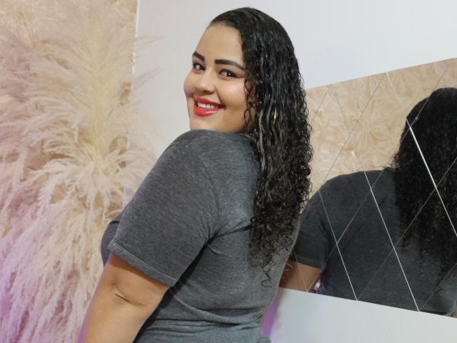 MelannieCarter with curly black hair, smiling, red lipstick, gray t-shirt, looking back over shoulder, reflection in mirror, puffy beige wall in background.