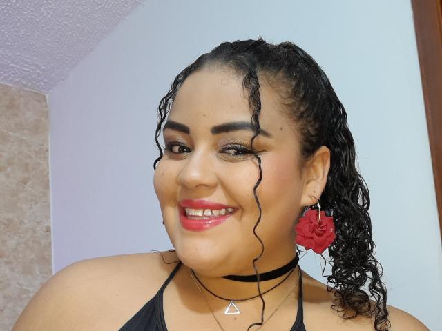 MelannieCarter heeft krullend zwart haar, rode lippenstift, een zwarte choker en rode oorbellen in de vorm van bloemen. Ze glimlacht, waarbij haar borsten licht zichtbaar zijn onder een zwarte top met halter. Ze is een schattige, mollige Latina..