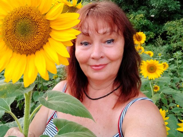 MilaMils with red hair, blue eyes, and striped dress, holding a giant yellow sunflower, smiling, wearing a black necklace, surrounded by more sunflowers.