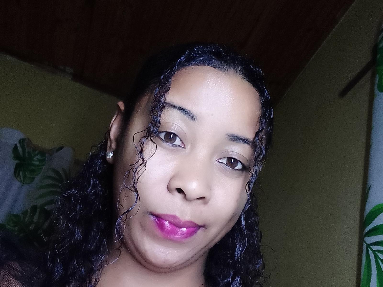 LaylahMorgane's face, brown eyes, curly black hair, pink lipstick, slight smile, light brown skin, wooden ceiling, green wall, white curtain with green leaves.