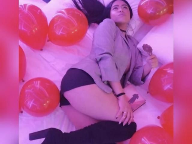 StellaBeckker sdraiata su un letto con palloncini rossi, indossa un blazer grigio, biancheria intima nera e stivali alti fino alla coscia, tiene in mano un sex toy, ha lunghi capelli neri e un braccialetto..