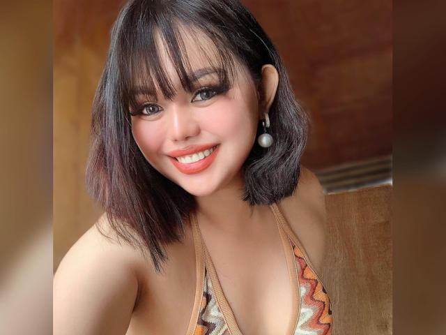 SweetAbbe with shoulder-length black hair, bangs, and a cute smile, wearing a patterned halter top, showing her small tits and wearing pearl earrings.