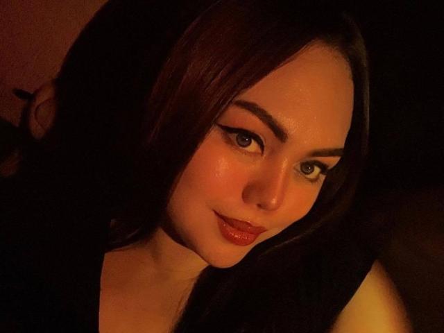 SweetAbbe with dark brown hair, full red lips, and thick black eyeliner, looking at the camera with a subtle smile in dim, warm lighting. Her ass is partially visible.