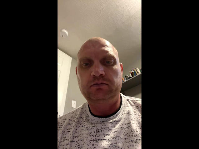 Bald Alexrus with a serious face, wearing a white and gray speckled shirt, has a slightly reddened neck and a small goatee, in a dimly lit room.