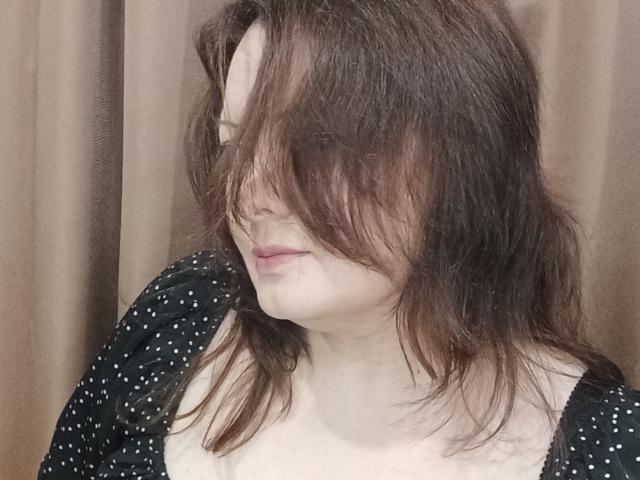 MayDreamy with messy brown hair covering her eyes, wearing a black polka dot top, has pale skin and a subtle, closed-lip smile. Her small, pale tits are partially visible.