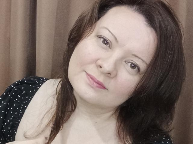 MayDreamy with pale skin, brown eyes, and pink lips, has medium brown, wavy hair, wearing a black, polka-dot top, slightly smiling, and looking to the side.