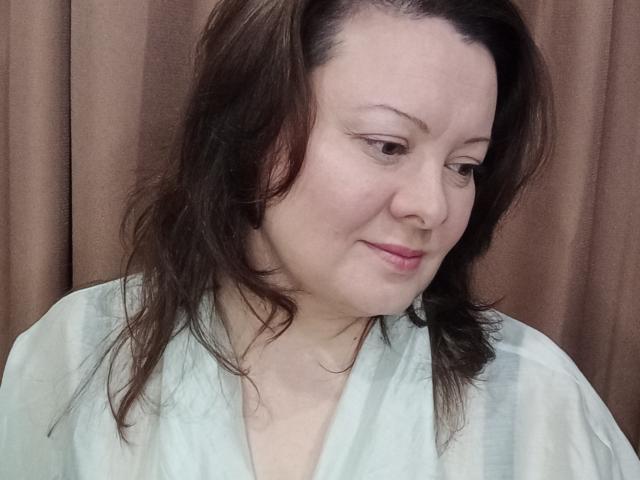 May, con la pelle chiara, capelli castani ondulati, un leggero rossetto rosa, indossa una veste verde, guarda in basso con un leggero sorriso, ha un anello al naso e tratti del viso delicati..