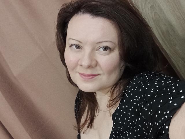 MayDreamy has pale skin, brown eyes, and brown hair, wearing a black polka dot top, showing her small boobs, with a slight smile and pink lipstick.