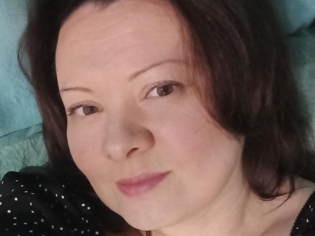 Il viso di MayDreamy, con la pelle chiara, gli occhi marroni e i capelli castano scuro ondulati, con un trucco leggero e un piccolo sorriso, indossa una maglietta nera a pois e giace sdraiata..