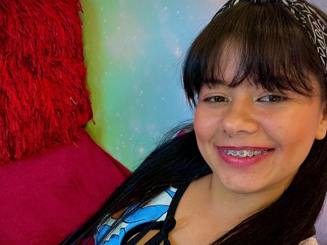 AprilDesire has dark brown hair with a black patterned headband, smiling with metal braces, wearing a blue and black patterned top, against a red and green background.