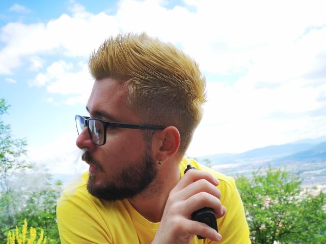 ChanceOfGod with yellow spiked hair, black glasses, beard, yellow shirt, holding a black camera, side profile, looking right, blue sky, white clouds, green trees in background.