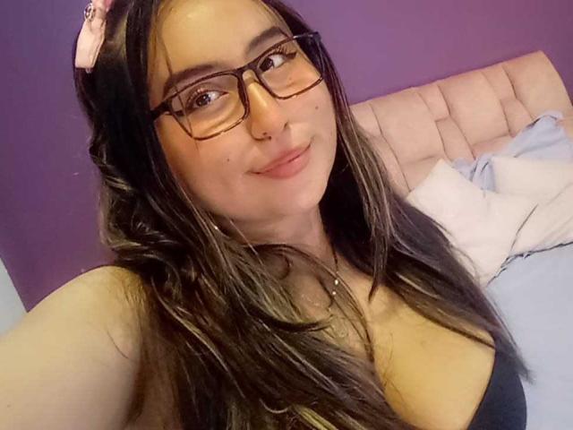 SalomettX, usando óculos, com um piercing no nariz e cabelos castanhos longos, sorrindo, mostrando o decote em uma blusa preta, deitada em uma cama com cabeceira rosa e parede roxa..