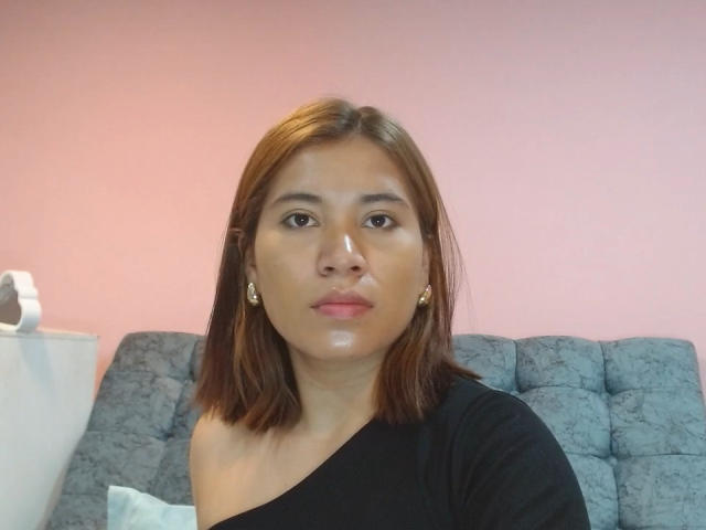 Bella, mit braunen, schulterlangen Haaren, einem schwarzen Top mit einem Schulterträger, goldenen Ohrringen und einem glänzenden Gesicht, sitzt auf einem grauen, gesteppten Sofa..