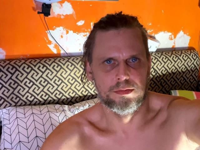OndraSekCZech has a shirtless, muscular upper body with a gray beard, short wet hair, and blue eyes, looking serious. Orange wall and patterned headboard in background.