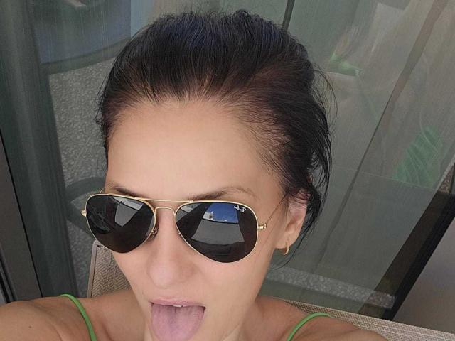 JoliDanielle with dark hair, tongue out, wearing gold aviator sunglasses, green spaghetti straps, and gold earrings, looking up. Her face is centered, with a slightly messy look.