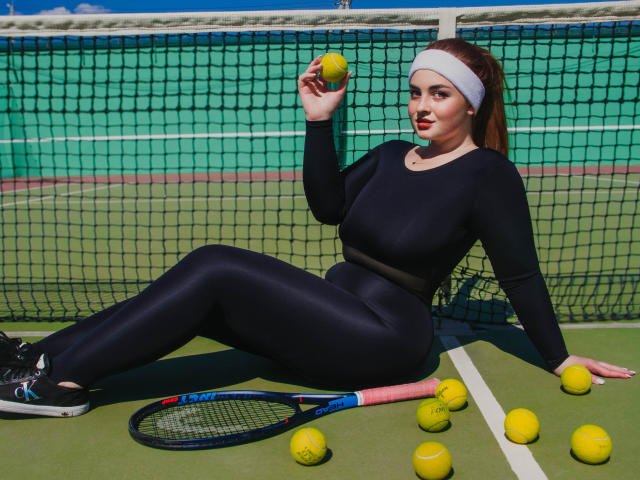 EsterMiln, with tits in a tight black sports outfit, sits on a tennis court, holding a yellow tennis ball, with a racket and more balls around her.