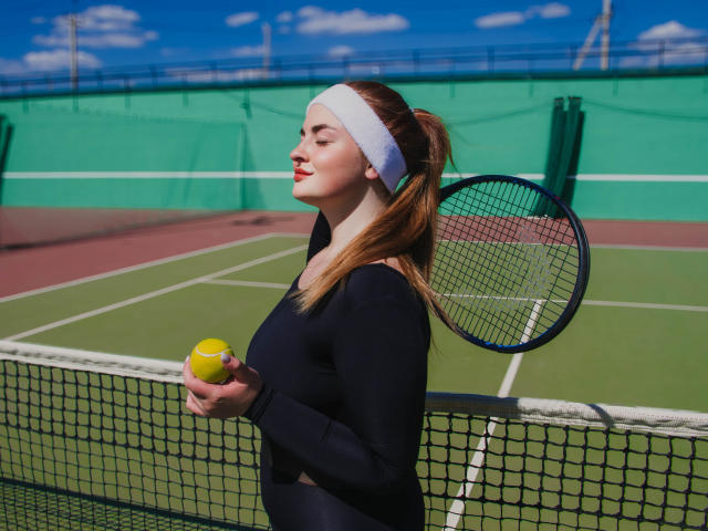 EsterMiln with a white headband, black long-sleeve top, holding a yellow tennis ball, and a tennis racket, standing near a net on a green tennis court.