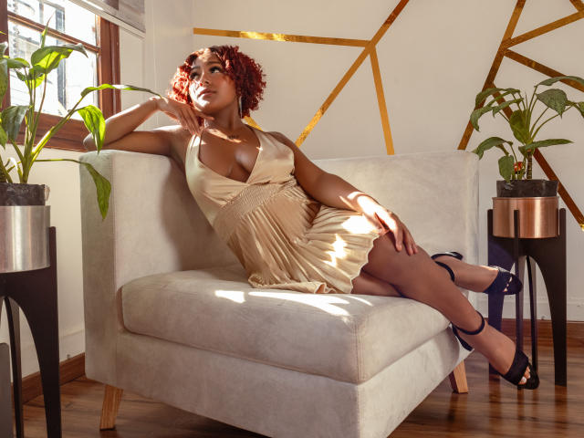 AmberQueeny, com cabelo ruivo encaracolado, seios grandes e pele escura, relaxando em um vestido bege, saltos pretos, em uma cadeira branca, com luz solar, plantas em vasos e detalhes dourados nas paredes..