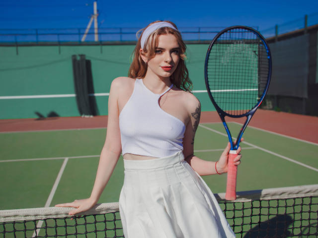 AdeleClifford with red hair, white sports bra, and high-waist white skirt, holding a blue and pink tennis racket, standing by a net on a sunny tennis court.