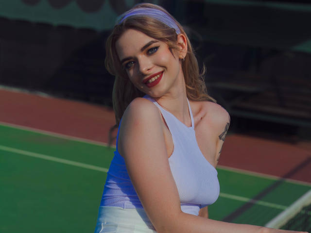 Adele Clifford, con cabello rubio, ojos azules y lápiz labial rojo, vestida con un top deportivo blanco y una diadema, sonriendo a la cámara, mostrando sus pequeños senos y un tatuaje en su brazo..