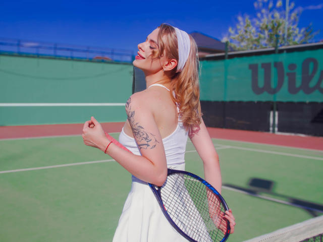 Adele Clifford, con cabello rubio, lleva una diadema blanca, una camiseta blanca y una falda blanca, sosteniendo una raqueta de tenis, mostrando un tatuaje en su brazo derecho, sonriendo, cielo azul..