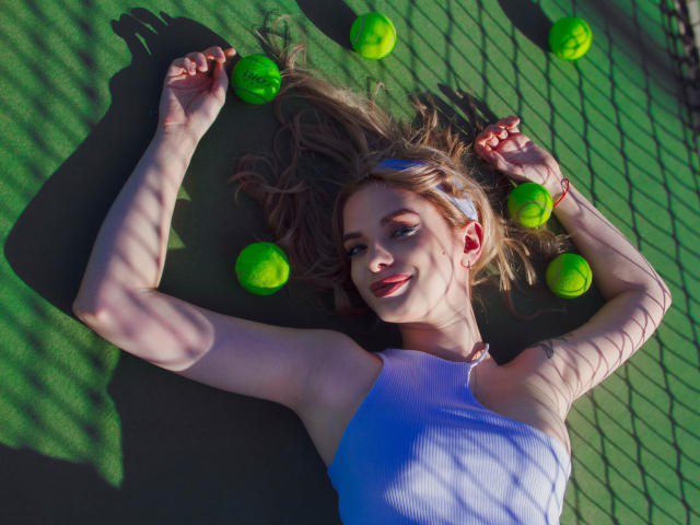 AdeleClifford acostada en la cancha de tenis verde, con los brazos por encima de la cabeza, vistiendo una camiseta blanca, rodeada de pelotas de tenis verdes, la luz del sol proyectando sombras, el cabello castaño largo extendido..
