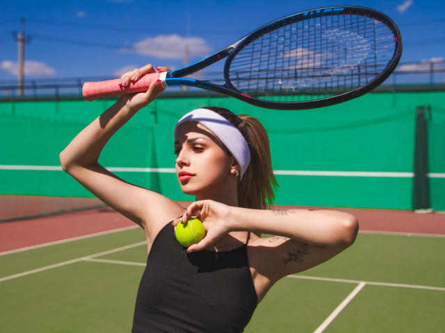 LaraMoss with a tennis racket, black sleeveless top, white headband, holding a green tennis ball, raising racket, sunny day, visible tattoo on right arm.