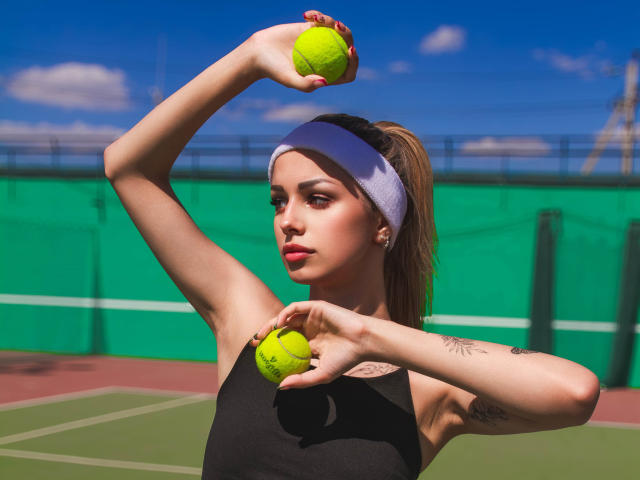 Lara Moss con una diadema morada, camiseta deportiva negra, sosteniendo pelotas de tenis amarillas en ambas manos, con los brazos levantados, tatuaje en el antebrazo derecho, fondo de cielo azul, día soleado..