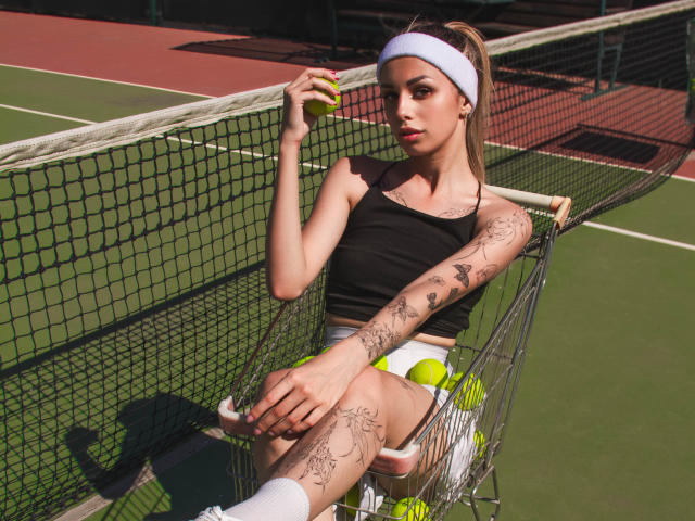 LaraMoss with tattoos, black tank top, white headband, tennis skirt, and socks, holding a yellow tennis ball, sitting on a net, tennis court, sunlight, relaxed, sexy.