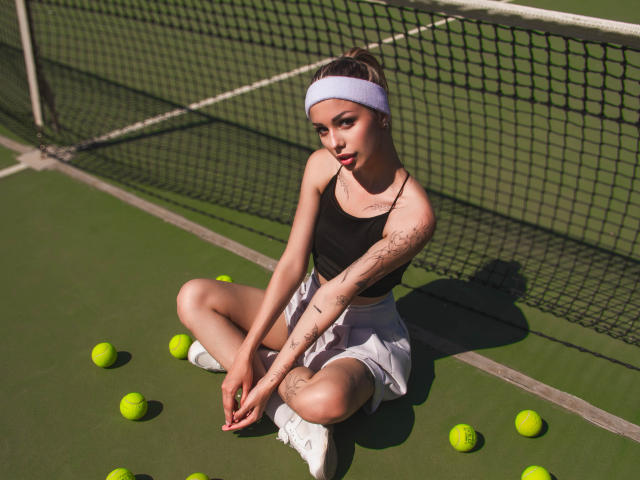 LaraMoss with tattoos, sitting on a tennis court, wearing a black top, white skirt, white sneakers, and a white headband, surrounded by tennis balls, looking at the camera.