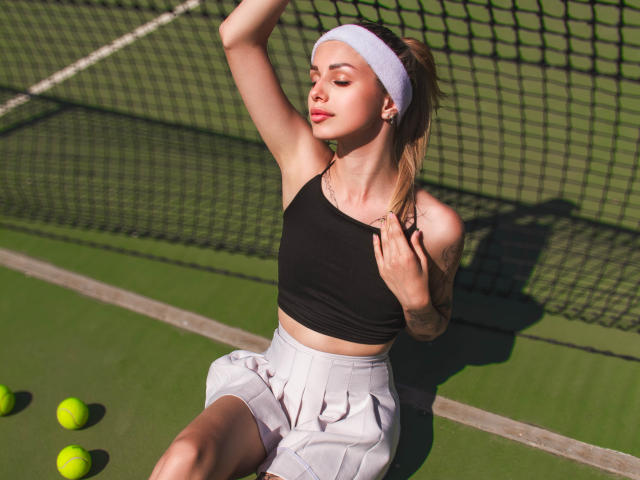 LaraMoss with arm raised, wearing black crop top, white headband, white pleated skirt, tattoos visible, sitting on green tennis court, yellow balls scattered, eyes closed, sunlit.