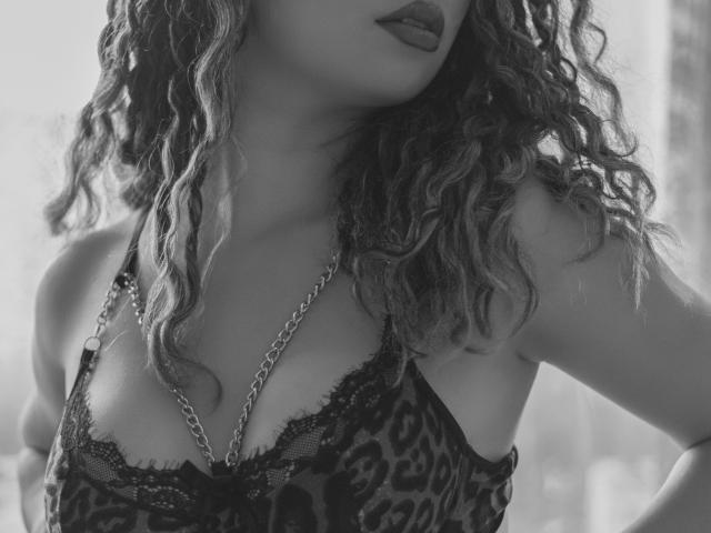 KendallPiierce69 with curly hair, wearing a leopard print lingerie top with lace, showcasing her tits in a black-and-white shot, chain necklace, and dark lipstick.